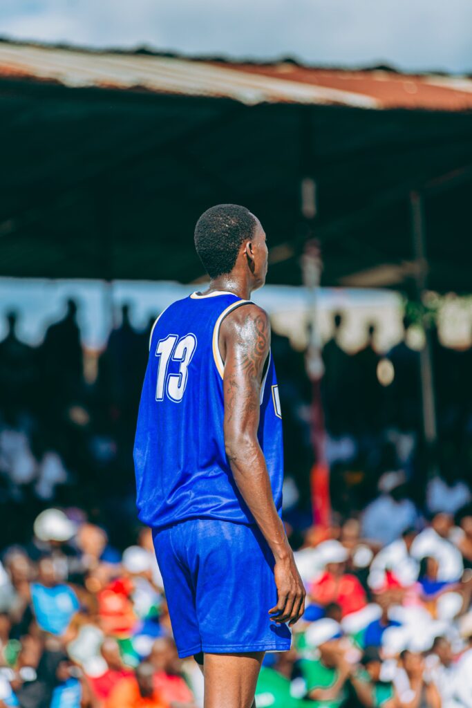 A male basketball player wearing a blue jersey with number 13 on a sunny outdoor court.